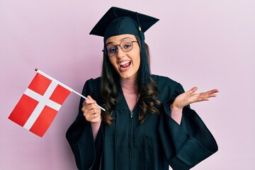 Young hispanic woman wearing graduation uniform holding denmark flag celebrating achievement with happy smile and winner expression with raised hand - obrazy, fototapety, plakaty