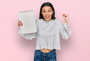 Young hispanic girl holding notebook screaming proud, celebrating victory and success very excited...