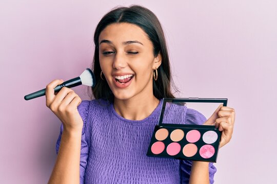 Young hispanic woman holding makeup brush and blush winking looking at the camera with sexy expression, cheerful and happy face.