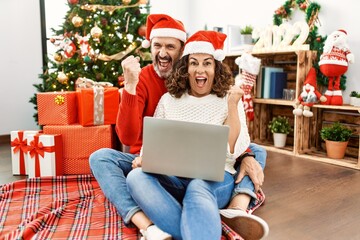 Obraz na płótnie Canvas Hispanic middle age woman and mature man using laptop sitting by christmas tree screaming proud, celebrating victory and success very excited with raised arms