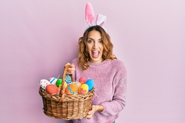 Young caucasian woman wearing cute easter bunny ears holding colored egg celebrating crazy and...