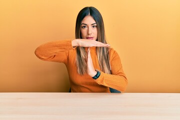 Young hispanic woman wearing casual clothes sitting on the table doing time out gesture with hands,...