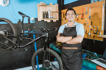 bicycle mechanic in smiling apron holding bicycle with crossed hands while standing beside bicycle in workshop