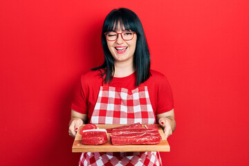 Young hispanic woman wearing cook apron holding board with meat winking looking at the camera with...
