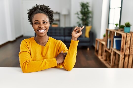 Young african american woman wearing casual clothes sitting on the table at home with a big smile on face, pointing with hand and finger to the side looking at the camera.
