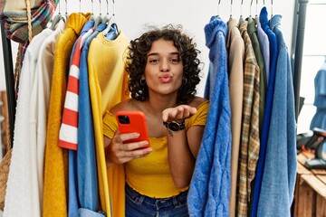 Young hispanic woman searching clothes on clothing rack using smartphone looking at the camera blowing a kiss with hand on air being lovely and sexy. love expression.