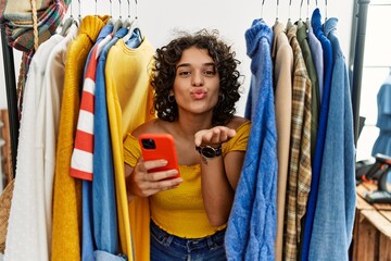 Fototapeta premium Young hispanic woman searching clothes on clothing rack using smartphone looking at the camera blowing a kiss with hand on air being lovely and sexy. love expression.