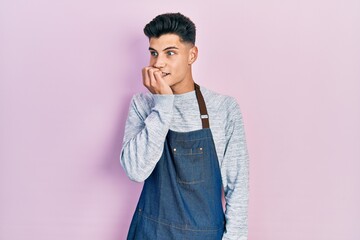 Fototapeta premium Young hispanic man wearing apron looking stressed and nervous with hands on mouth biting nails. anxiety problem.