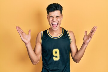 Young hispanic man wearing basketball uniform celebrating mad and crazy for success with arms raised and closed eyes screaming excited. winner concept