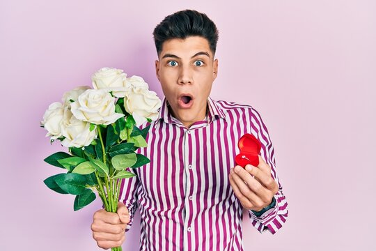 Young hispanic man holding bouquet of flowers and wedding ring afraid and shocked with surprise and amazed expression, fear and excited face.