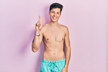 Fototapeta premium Young hispanic man wearing swimwear shirtless smiling with happy face winking at the camera doing victory sign. number two.