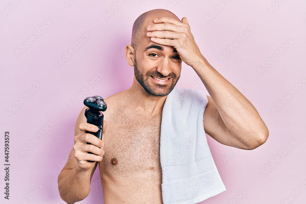 Poster young bald man shirtless holding electric razor stressed and frustrated with hand on head, surprised