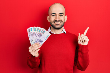 Young bald man holding egyptian pounds banknotes smiling happy pointing with hand and finger to the side