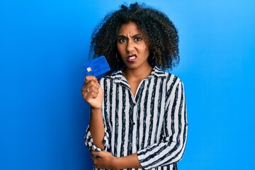 Beautiful african american woman with afro hair holding credit card clueless and confused expression. doubt concept.