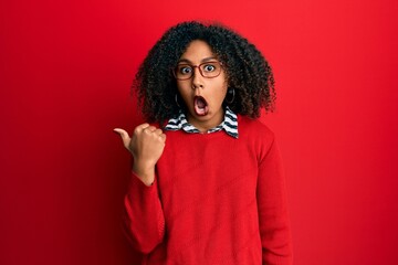 Fototapeta na wymiar Beautiful african american woman with afro hair wearing sweater and glasses surprised pointing with hand finger to the side, open mouth amazed expression.