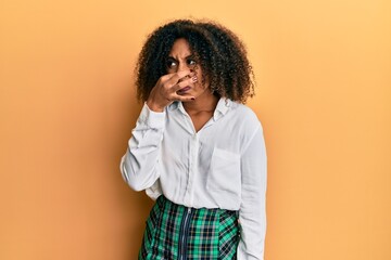 Beautiful african american woman with afro hair wearing scholar skirt smelling something stinky and disgusting, intolerable smell, holding breath with fingers on nose. bad smell
