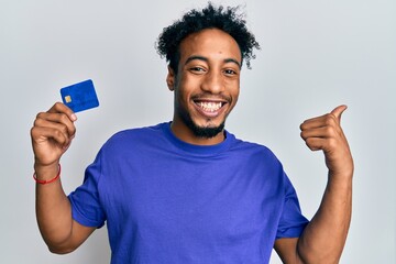 Young african american man with beard holding credit card pointing thumb up to the side smiling happy with open mouth