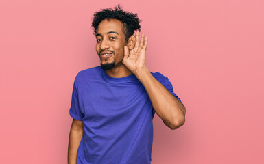 Young african american man with beard wearing casual purple t shirt smiling with hand over ear listening an hearing to rumor or gossip. deafness concept.