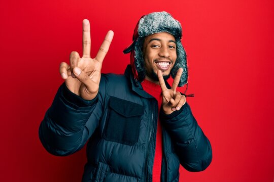 Young african american man with beard wearing winter hat and coat smiling looking to the camera showing fingers doing victory sign. number two.