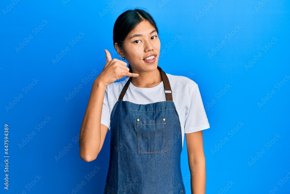 Poster Young chinese woman wearing waiter apron smiling doing phone gesture with hand and fingers like talking on the telephone. communicating concepts.