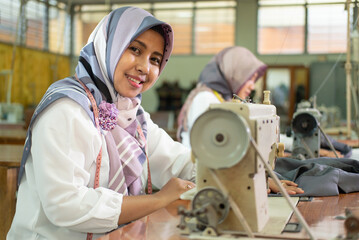 Muslim female employees wearing headscarves smile while working using sewing machines in the...