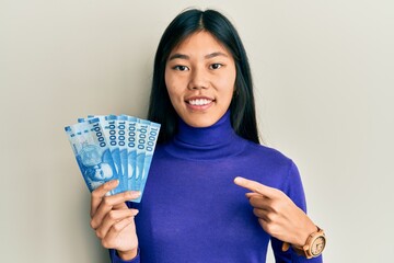 Young chinese woman holding 10000 chilean pesos smiling happy pointing with hand and finger