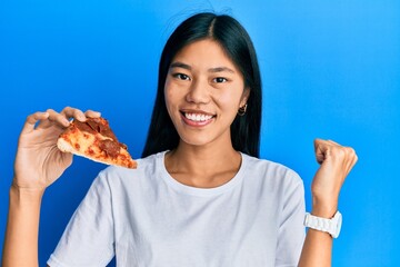 Young chinese woman eating tasty pepperoni pizza screaming proud, celebrating victory and success very excited with raised arms