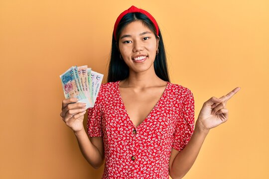 Young chinese woman holding singapore dollars banknotes smiling happy pointing with hand and finger to the side