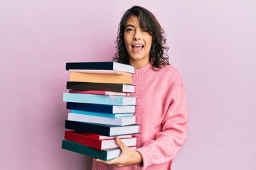 Young hispanic woman holding a pile of books winking looking at the camera with sexy expression, cheerful and happy face.