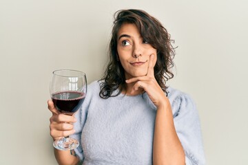Young hispanic woman drinking a glass of red wine serious face thinking about question with hand on...