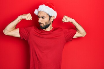 Young hispanic man wearing christmas hat showing arms muscles smiling proud. fitness concept.