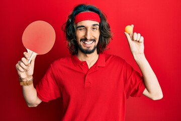 Young hispanic man holding red ping pong racket and ball winking looking at the camera with sexy expression, cheerful and happy face.