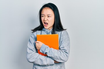Young chinese girl holding book angry and mad screaming frustrated and furious, shouting with anger. rage and aggressive concept.