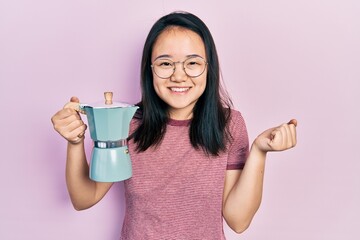 Young chinese girl holding italian coffee maker screaming proud, celebrating victory and success very excited with raised arm