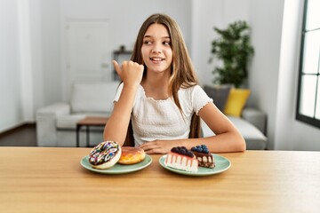 Young brunette teenager eating cakes and sweets pointing thumb up to the side smiling happy with open mouth