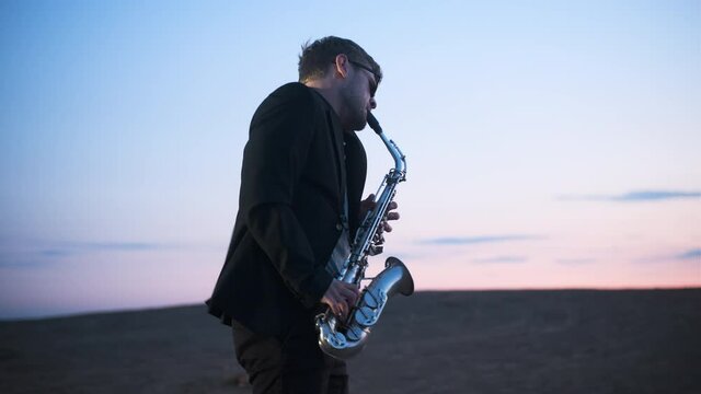 bearded musician in sunglasses and black jacket performs his part on beautiful shiny saxophone at sunset or sunrise with blue-pink sky in the background. Jazz play nature.