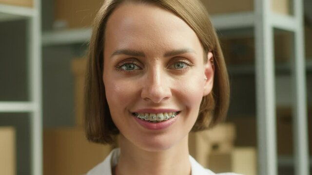Young smiling woman sitting in storage close-up, smile with braces of female employee of warehouse, looking in camera on background of cardboard boxes. Delivering and logistics, small woman business.