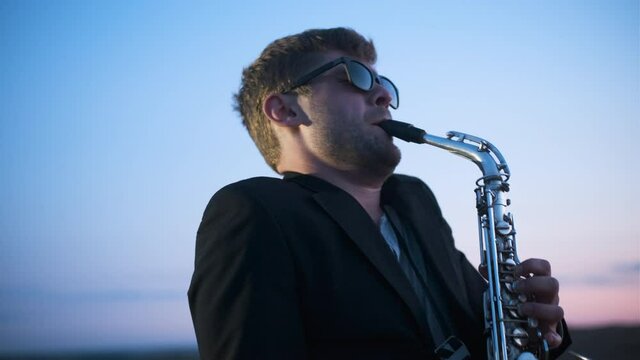 Portrait of bearded musician in sunglasses and black jacket performs his part on beautiful shiny saxophone at sunset or sunrise with blue-pink sky in the background. Jazz play nature.