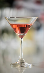Close up vertical shot of tall martini cocktail glass with red pink alcoholic drink beverage on counter bar table at celebration party night in pub in front of blurred background environment