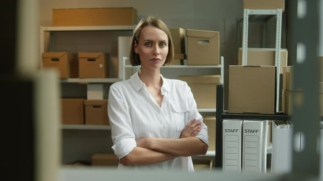 Young smiling woman standing in storage, female employee of warehouse wearing formal office clothes, looking in camera on background of cardboard boxes. Delivering and logistics, small woman business.