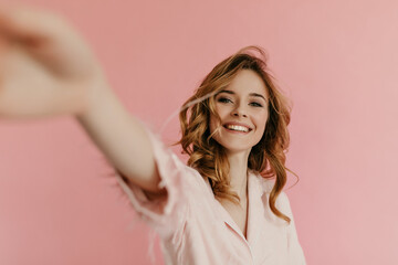 Cheerful curly woman in pink outfit makes photo on isolated background. Wavy haired lovely lady with cute smile takes salfie..