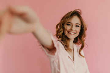 Smiling curly woman in pink outfit takes selfie on isolated background. Stylish girl in light pajamas looking into camera..