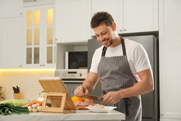 Man cutting chicken fillet while watching online cooking course via tablet in kitchen