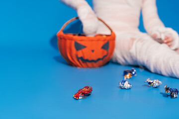 Child in mummy costume eats candies from basket in the form of pumpkin against blue background Halloween trick or treat
