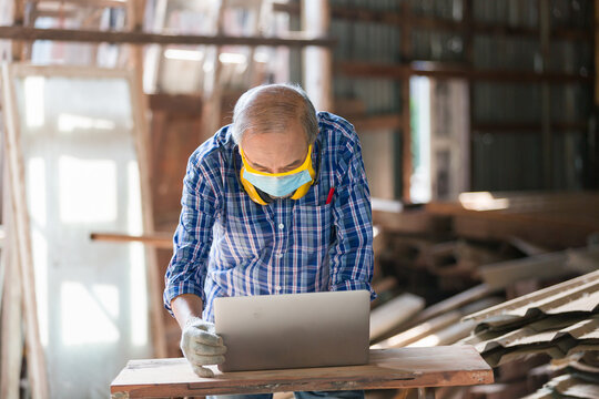 Asian senior male carpenter using computer laptop and wearing safety goggles and mask at wood shop. Woodworker working with computer laptop. Male carpenter working with computer laptop - Powered by Adobe