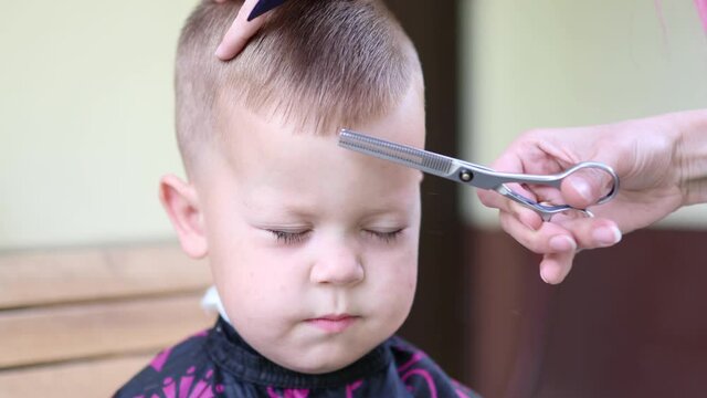 very close-up view of front portrait of a little baby boy with closed eyes while getting haircut with scissors. hairdresser's nah in work