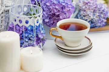 A visual for content. Still life in vintage style. A mug with a drink, an old book, a cage, candles and hydrangea flowers in the garden on a white wooden table. The concept of a tea ceremony.