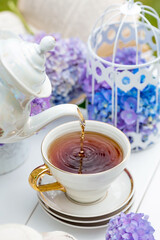 A visual for content. Still life in vintage style. A mug with a drink, an old book, a cage, candles and hydrangea flowers in the garden on a white wooden table. The concept of a tea ceremony.