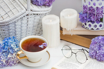 A visual for content. Still life in vintage style. A mug with a drink, an old book, a cage, candles and hydrangea flowers in the garden on a white wooden table. The concept of a tea ceremony.