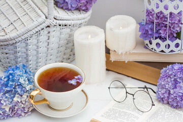 A visual for content. Still life in vintage style. A mug with a drink, an old book, a cage, candles and hydrangea flowers in the garden on a white wooden table. The concept of a tea ceremony.